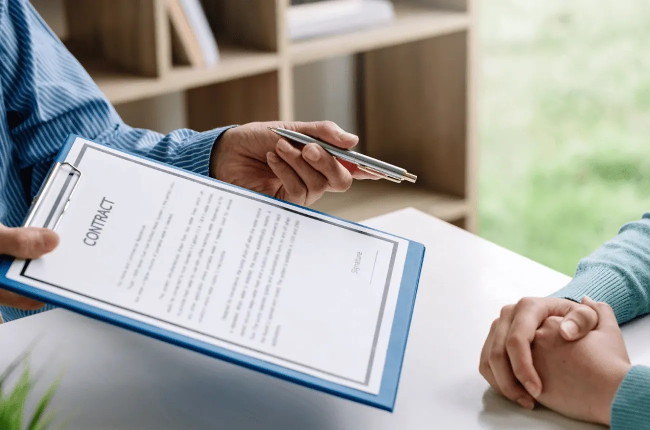 A person holding out a contract on a blue clipboard and a pen to someone sitting across a white desk