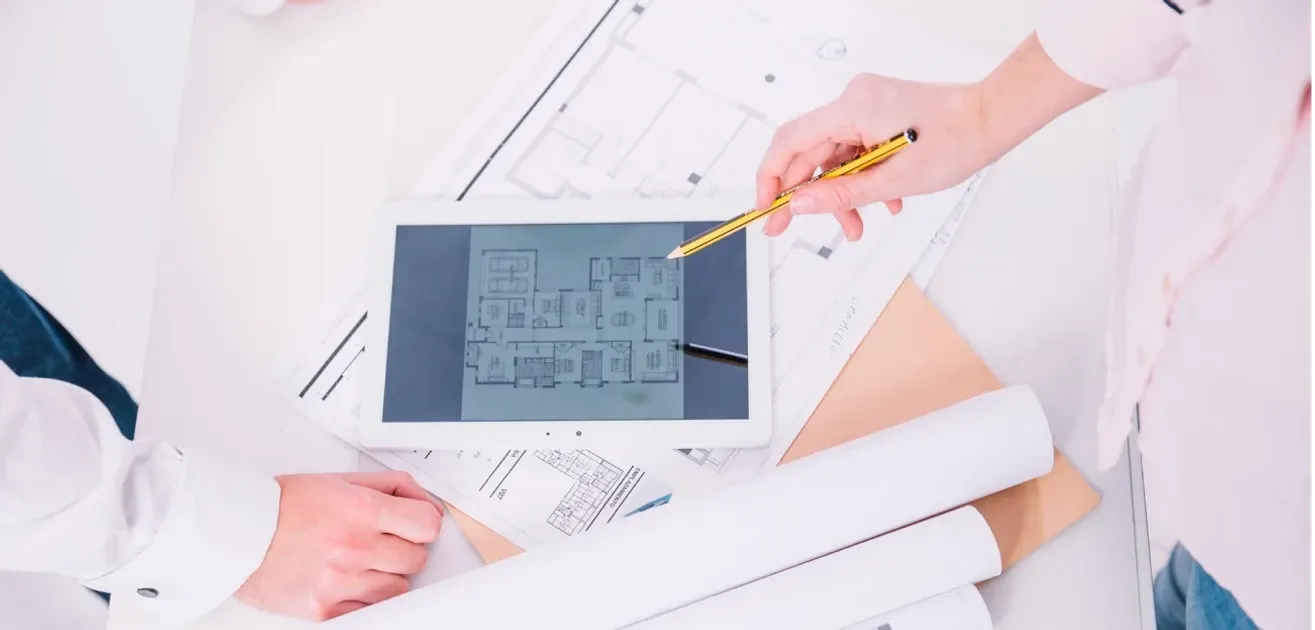 A hand pointing a pencil at a house floor plan on a tablet, lying on printed blueprints and rolled drawings