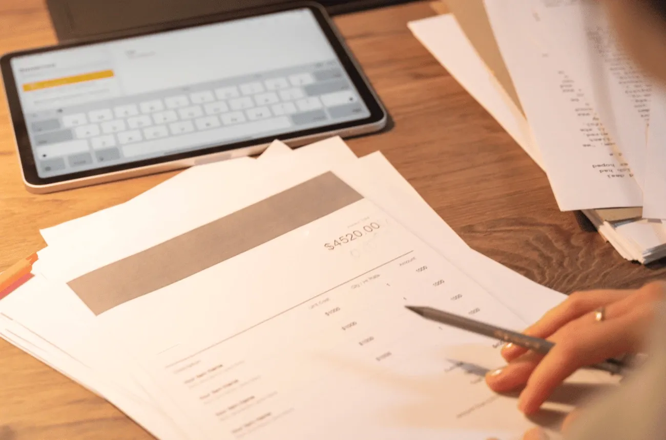 A hand holding a pencil over a printed invoice on a wooden desk next to a tablet and a stack of papers