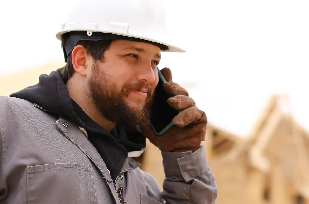 A bearded worker in a white hard hat and work gloves talking on a phone in front of a house frame under construction