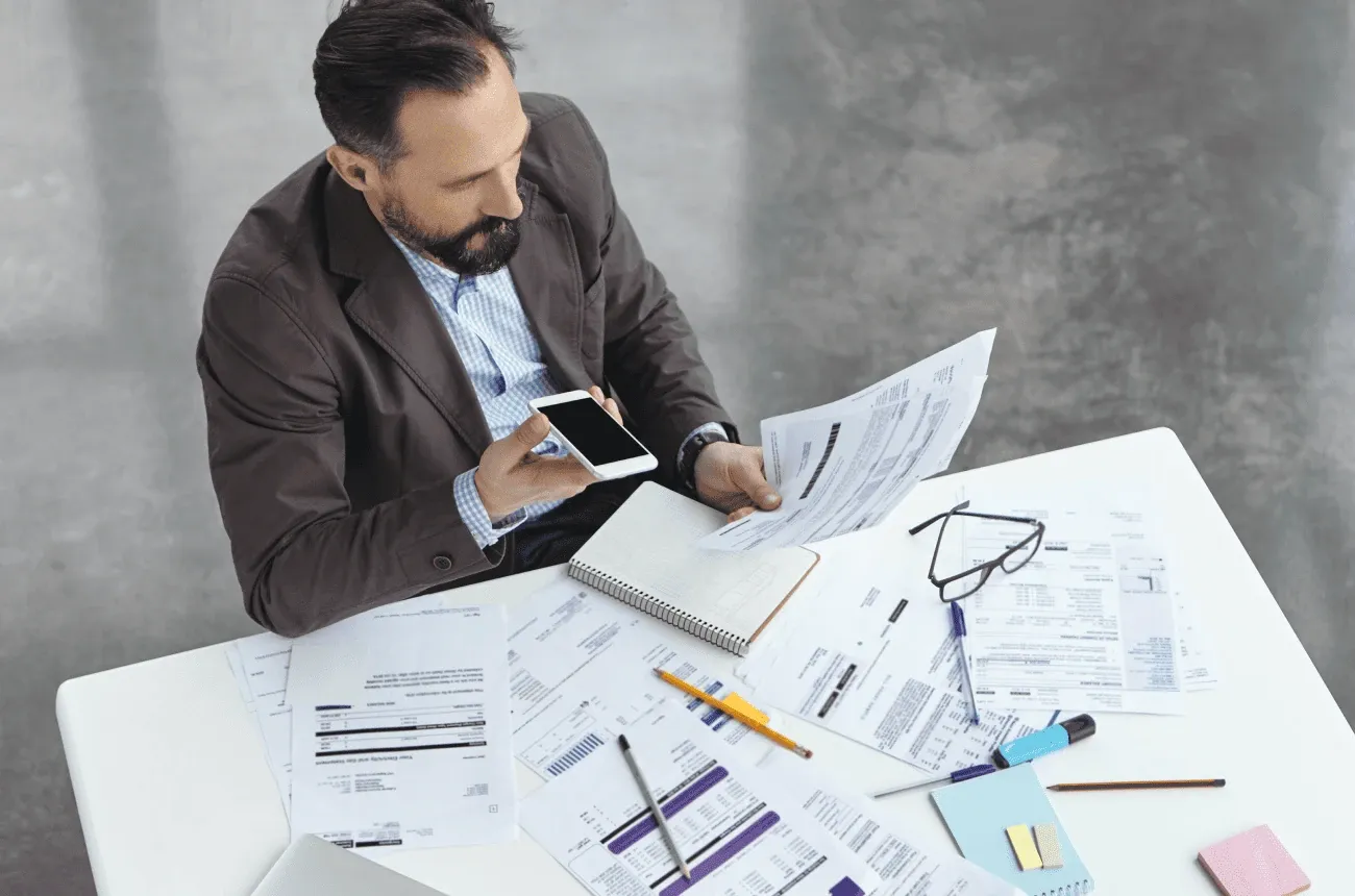 A man in a blazer holds a phone and papers at a white desk covered in printed bills and invoices